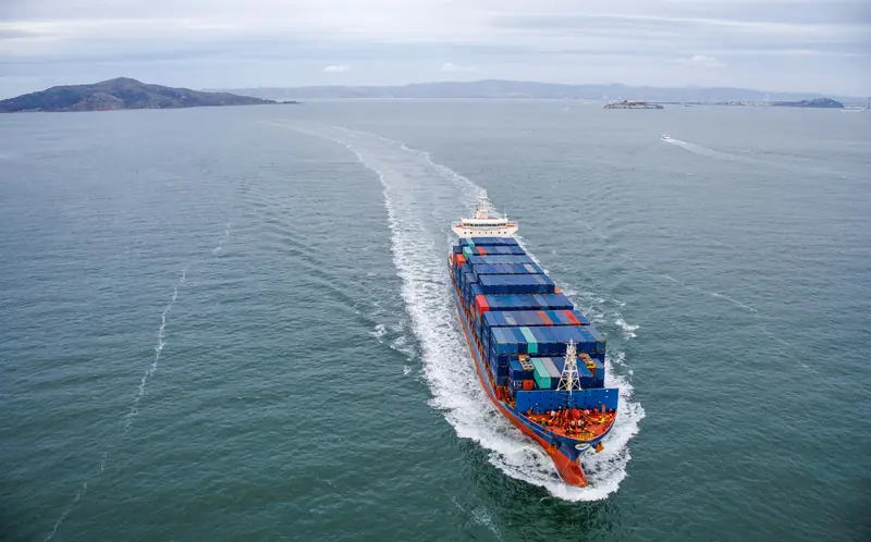 Porte-conteneurs en route, vu du ciel, sillage blanc sur une mer calme et collines à l'horizon